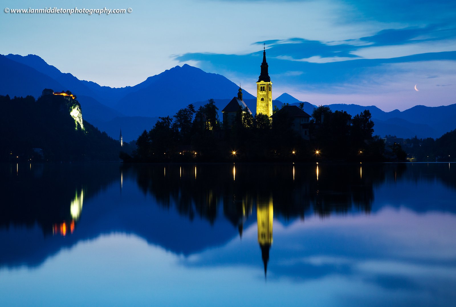 View across the beautiful Lake Bled, island church of the assumption of Mary, and hilltop castle, all topped off with the crescent of the waning moon at dawn, Slovenia.