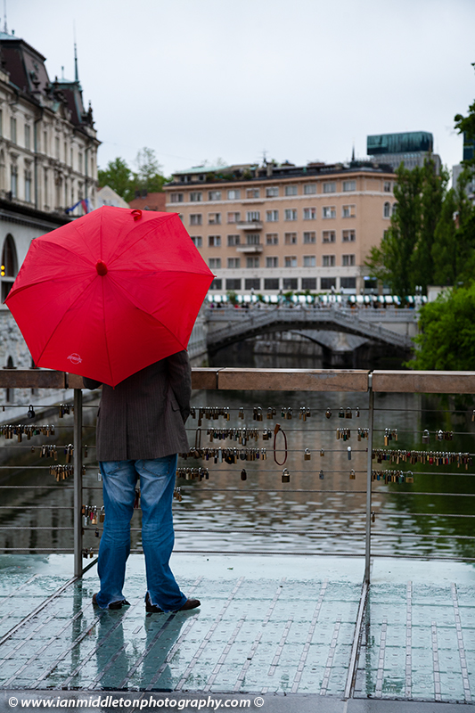 Man holding a red umbrella and standing on The Butchers' Bridge looking over the Ljubljanica river towards the Trznica (market) Triple Bridge on a rainy day in Ljubljana, Slovenia. The wire fence on the bridge is covered with padlocks put there by locals and tourists. All this region was designed by Slovenia's most celebrated architect, Joze Plecnik.